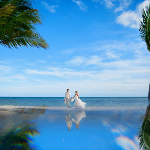 A soon-to-be married couple walks down the beach between two palm trees.